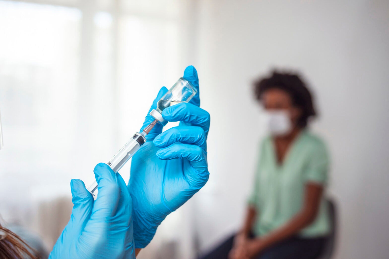 A doctor wearing gloves filling a syringe with a vaccine for COVID-19 while a patient sits in the background.