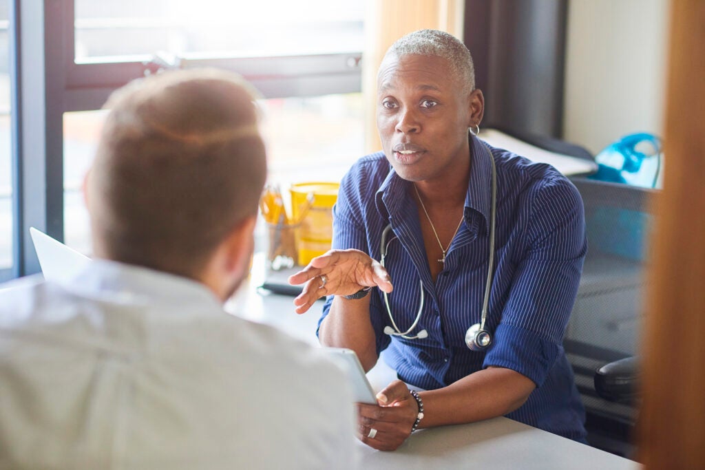 A female doctor sits at her desk and chats to a male patient while looking at his test results on her digital tablet .  She wears a blue shirt with the sleeves rolled up and a stethoscope around her neck.